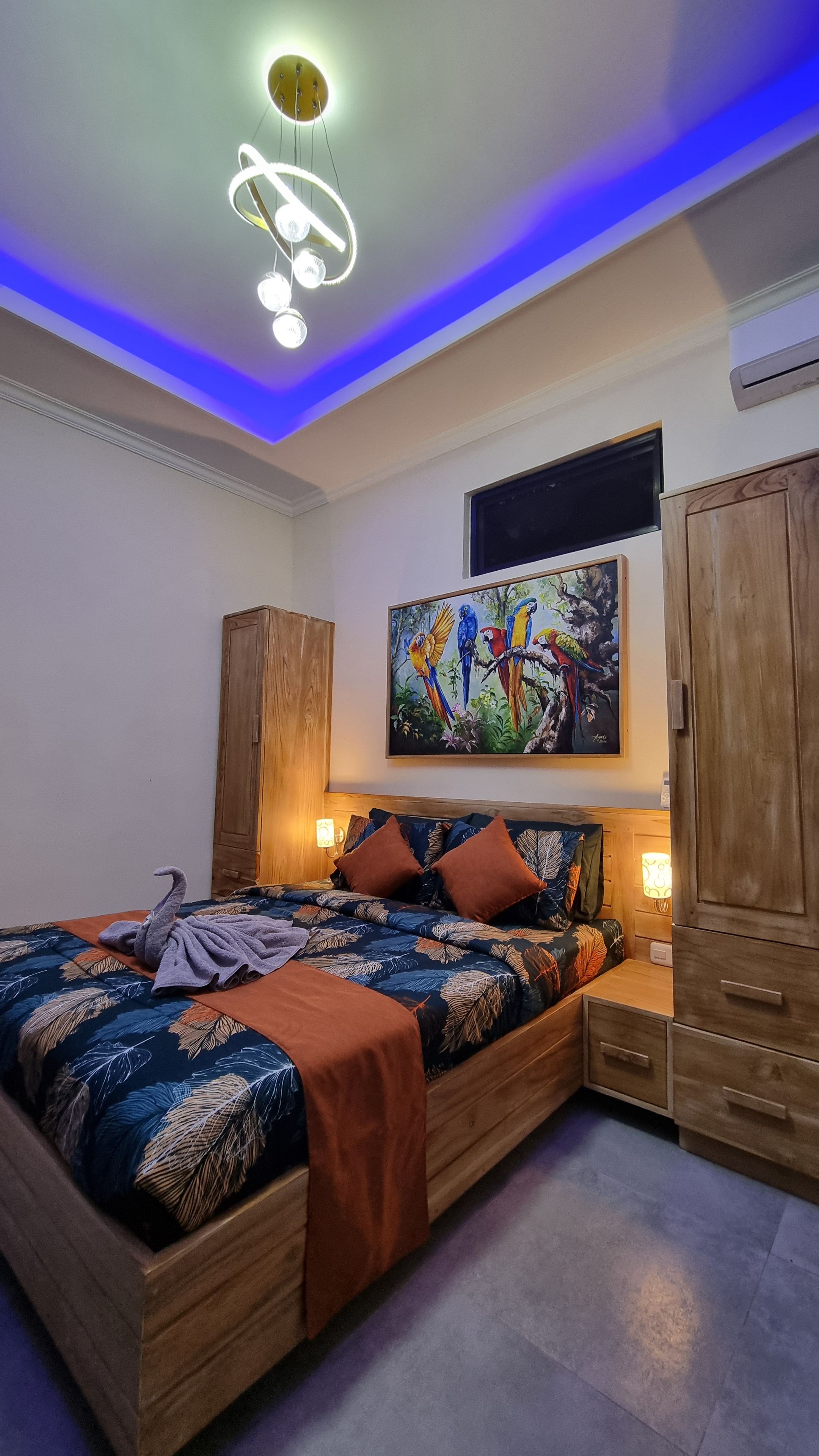 A bedroom with a bed, a wooden wardrobe, a painting of parrots, and a ceiling light fixture.