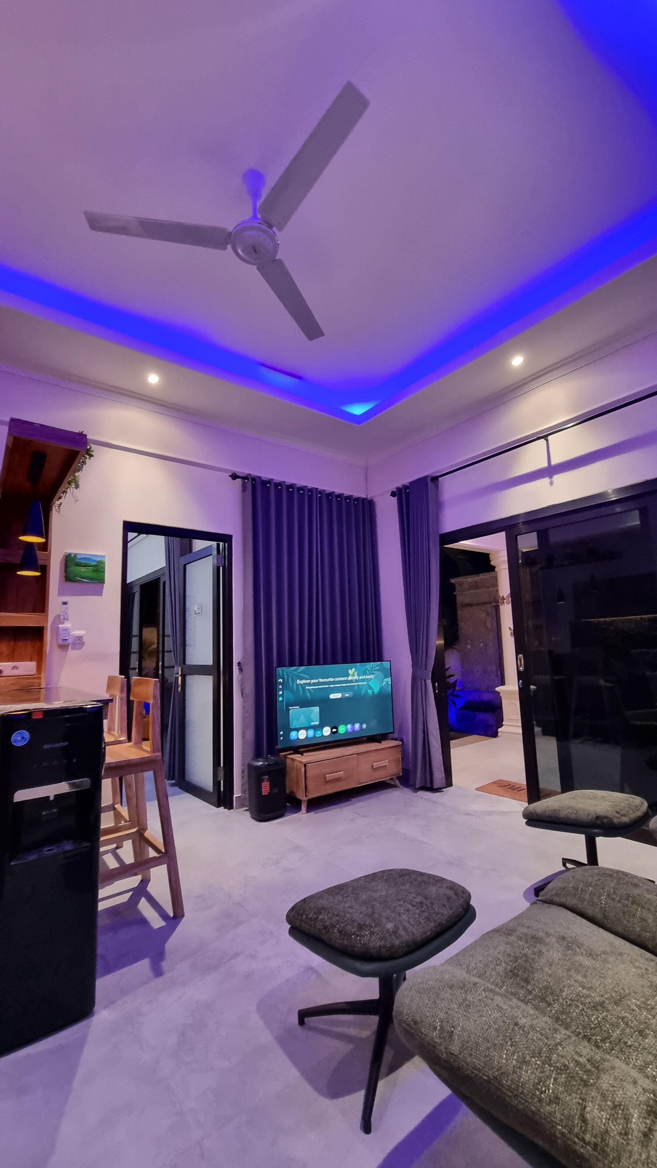 Una sala de estar moderna con ventilador de techo, televisor de pantalla plana y una zona de estar con sofá y reposapiés.