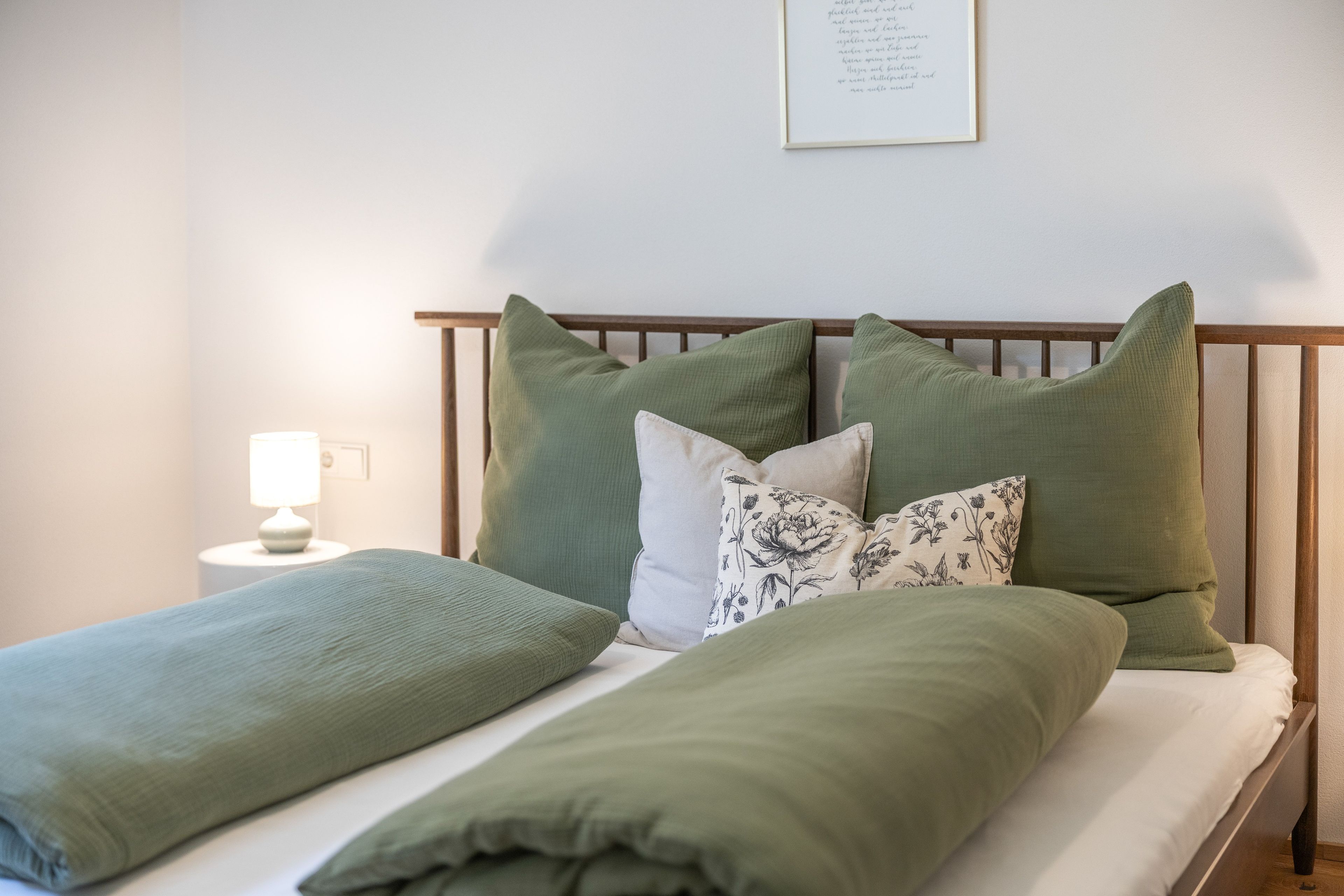 Una cama bien tendida con ropa de cama verde y un cojín floral, una lámpara de noche y un texto enmarcado en la pared.