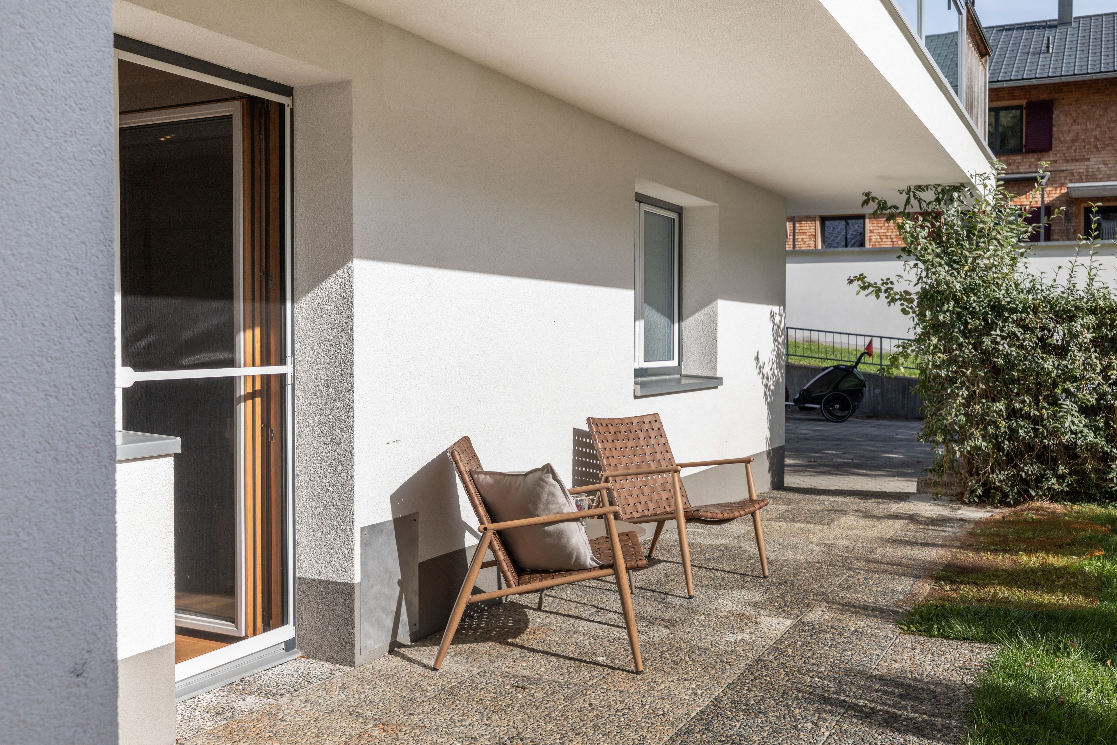 A patio with two wicker chairs and a small table, a sliding glass door, and a view of a street with a parked car and a building in the background.