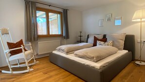 Un dormitorio con cama, una mecedora, una ventana con cortinas, un radiador y una lámpara.