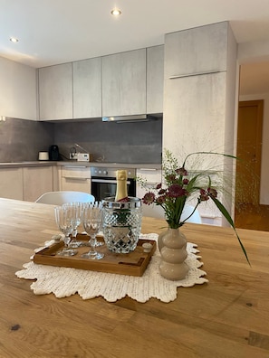 A modern kitchen with a wooden table set for a celebration, featuring a champagne bottle, glasses, and a vase with flowers.