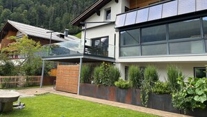 Una casa moderna de dos pisos con un balcón, una terraza de madera y un jardín con diversas plantas.