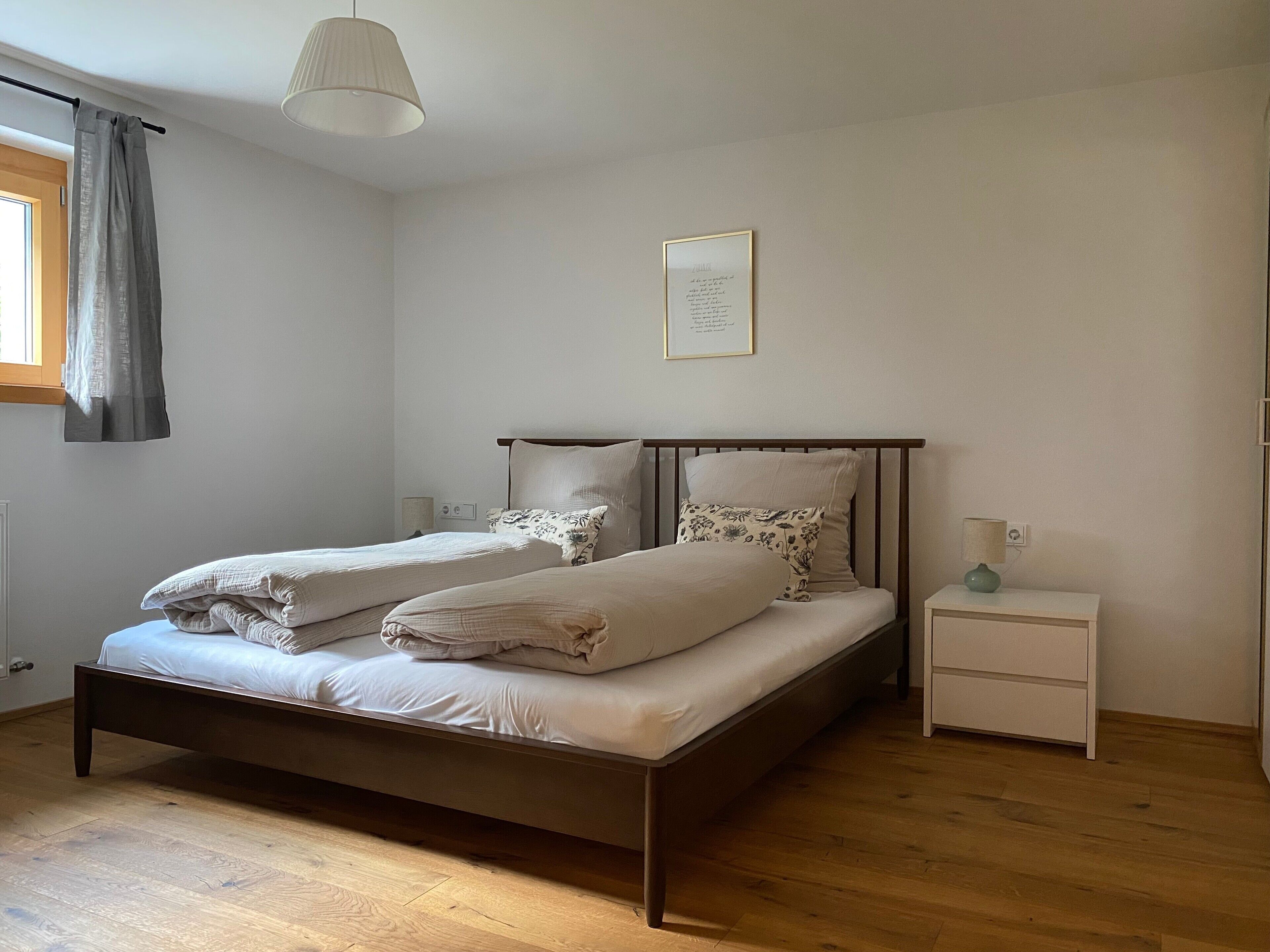 Un letto matrimoniale con struttura in legno e due cuscini, un comodino con una lampada, e una finestra con tenda.
