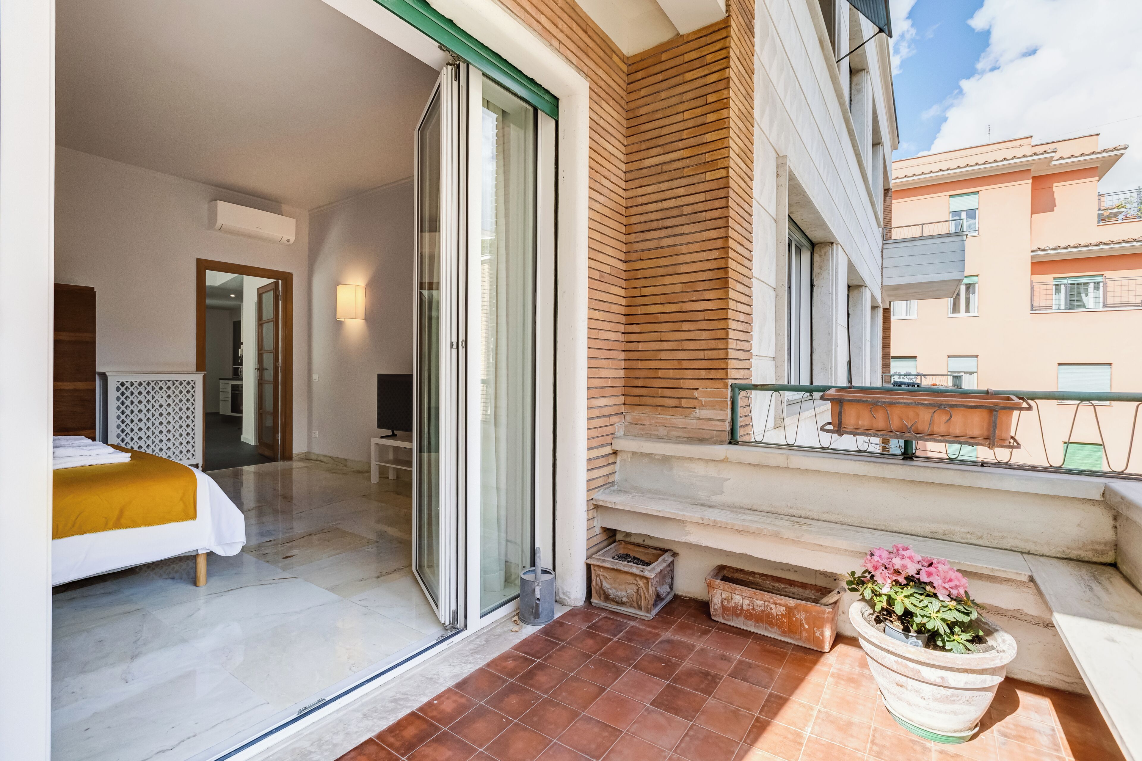 A hotel room with a bed, a sliding glass door, a balcony with a potted plant, and a view of neighboring buildings.