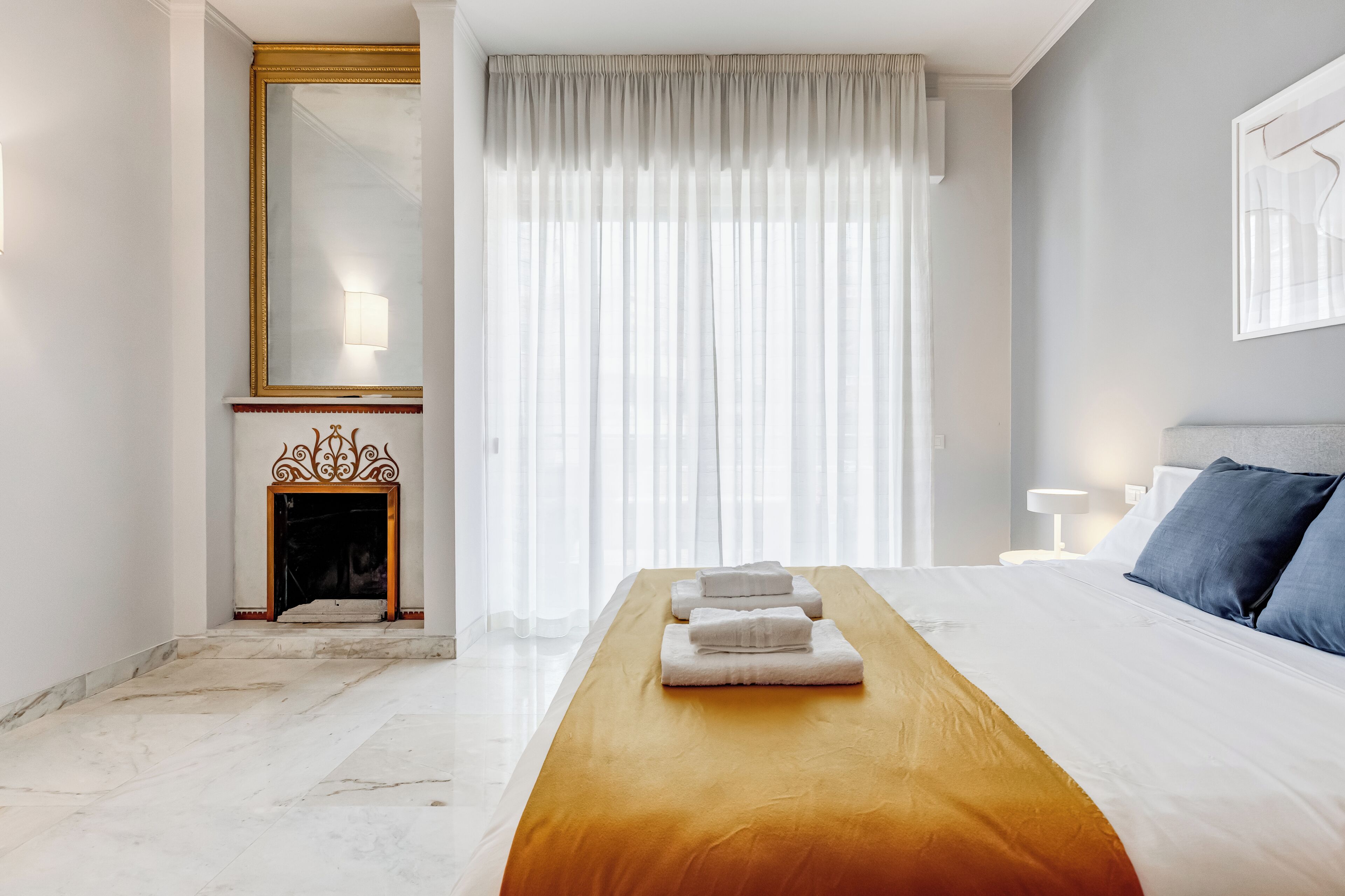 A hotel room with a bed, a wooden bench, a fireplace, and a large window with curtains.