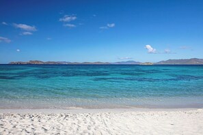 Ein Strand mit klarem türkisblauem Wasser, weißem Sand und in der Ferne erkennbaren Landformen unter blauem Himmel.