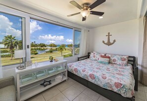 Ein Schlafzimmer mit einem Bett, einem Deckenventilator, einem Fenster mit Blick auf den Strand und einer Wanddekoration als Anker.