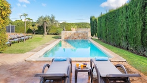 Una piscina rectangular con una pared de piedra en el centro, rodeada por un césped cuidado y un seto alto.