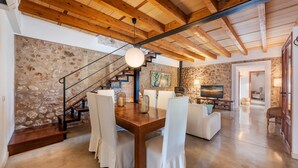 Amplia sala de estar con techo de madera, paredes de piedra, una escalera y una mesa de comedor con sillas.