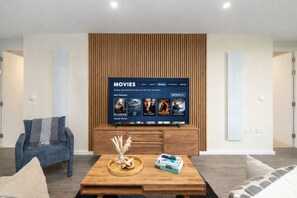 Ein modernes Wohnzimmer mit einem Flachbildfernseher, der ein Filmangebot zeigt, einem Couchtisch aus Holz mit Dekorationen und einem blauen Sessel.