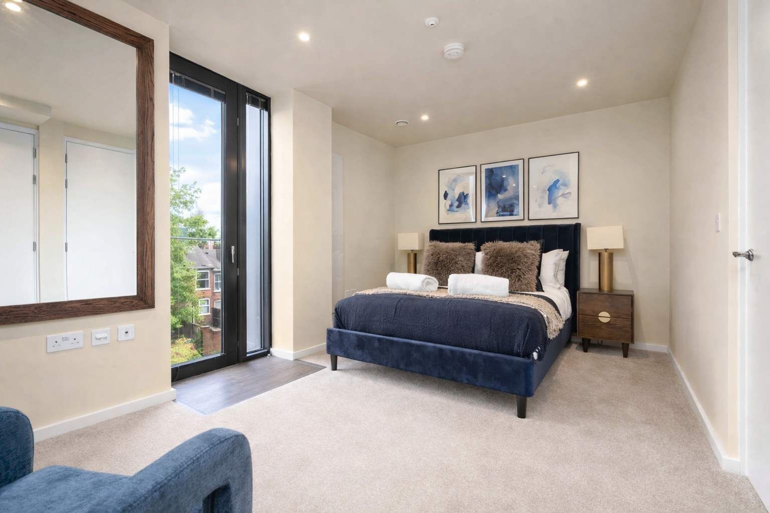 A bedroom with a large bed, a nightstand, a mirror, and a view of the outdoors.