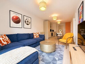Ein modernes Wohnzimmer mit einem blauen Sofa, einem gelben Sessel und einem Couchtisch. An der Wand befinden sich gerahmte Sportbälle und im Hintergrund ist eine Küche zu sehen.