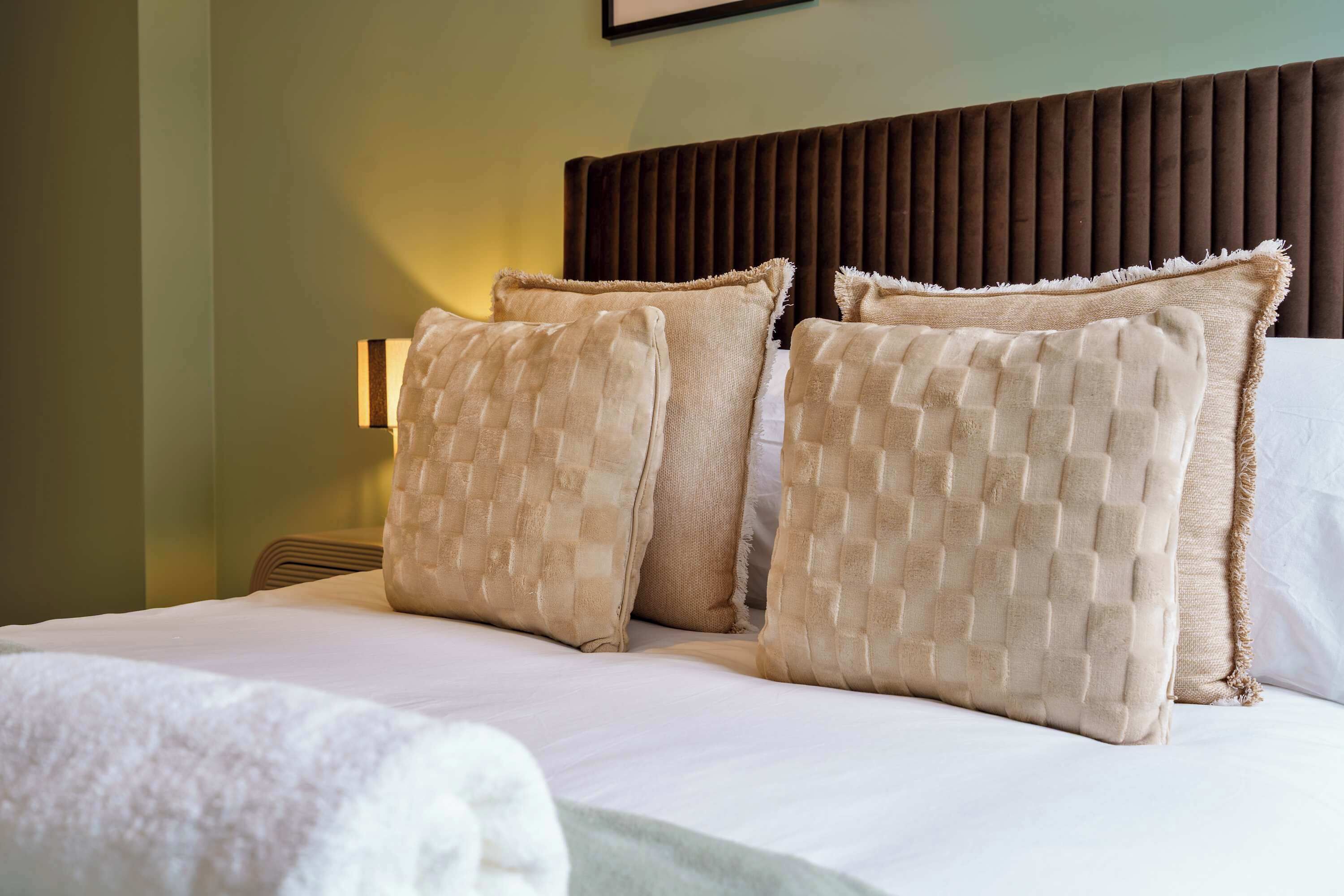 Quarto de hotel com cama, duas almofadas, cabeceira e uma luminária fixada na parede.