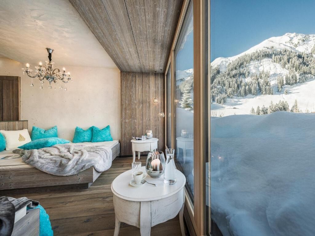 A bedroom with a bed, a chandelier, a small round table with a coffee pot, and a view of a snowy mountain landscape.