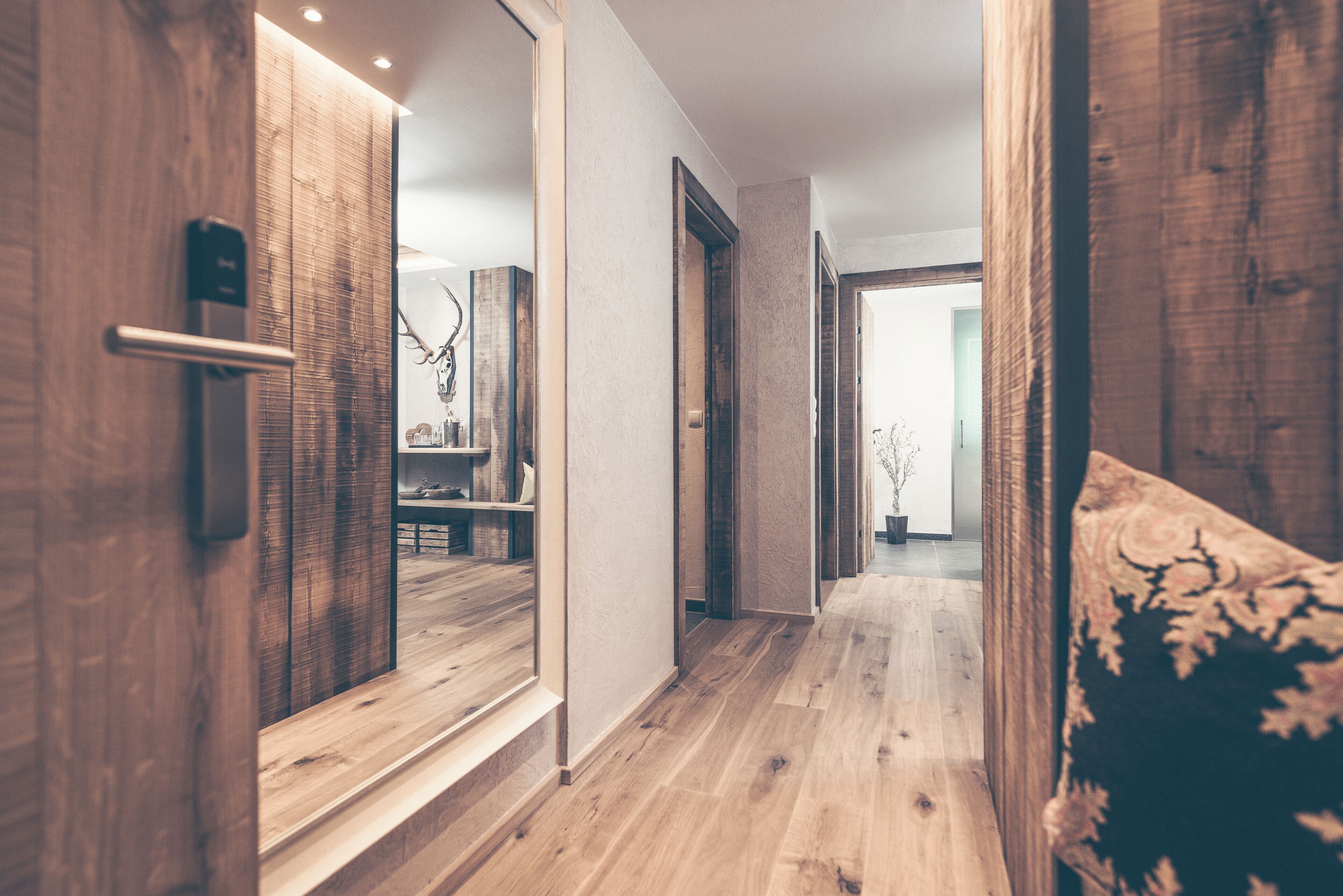 A hallway with wooden flooring, a mirror, and a door leading to a room with a bed and a decorative wall piece.