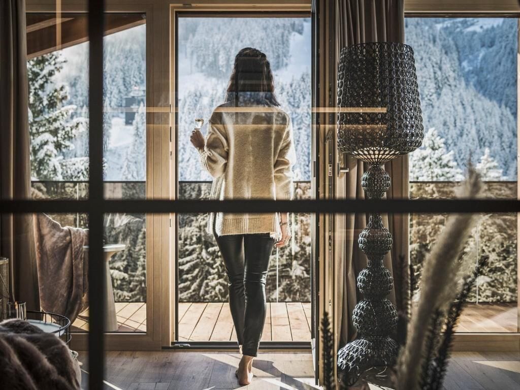 A person standing by a large window with a view of snow-covered trees and mountains.
