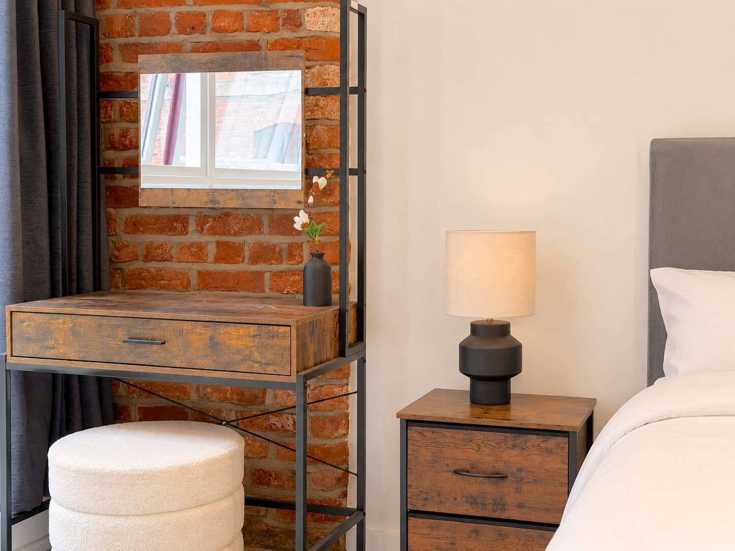 Ein Hotelzimmer mit Backsteinwand, einem hölzernen Schreibtisch mit Schubladen, einem Nachttisch mit Lampe, einem weißen Pouf und einem Bett mit weißer Bettwäsche.