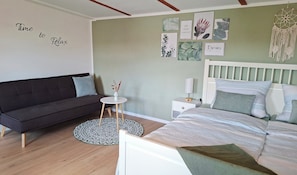 A modern bedroom with a dark sofa, a round side table, a bed with a white headboard, and wall art. - New, incredibly cozy vacation home in an old barn, near Lake Ellertshäuser (Schonungen)
