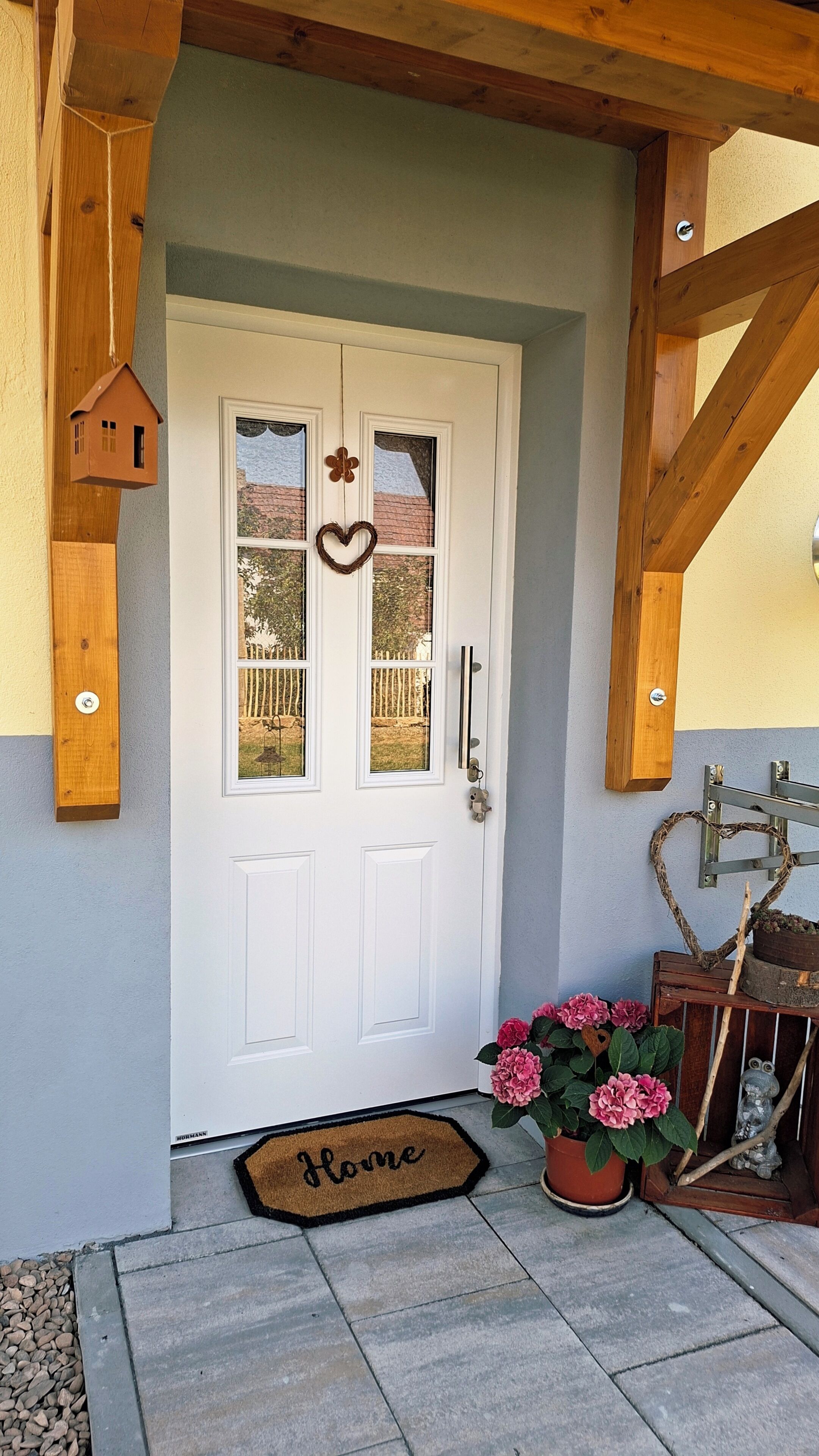 Une porte blanche ornée d’un cœur, un petit objet décoratif en forme de maison en bois, et un paillasson sur lequel est écrit le mot « Accueil ».