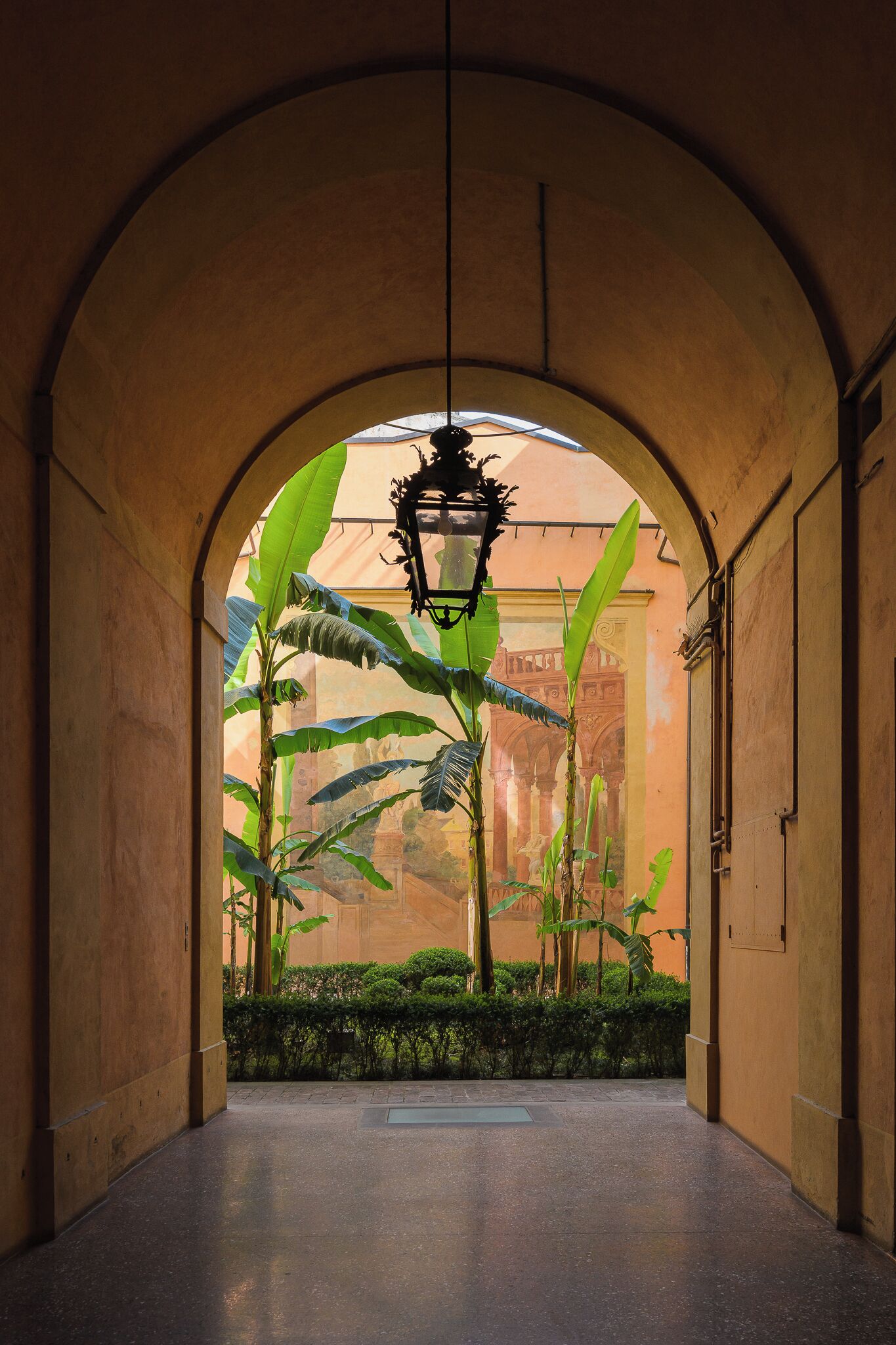 An arched corridor with a hanging lantern, a large window, and a view of a building with arches.