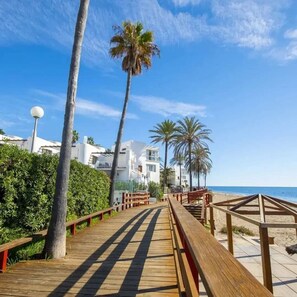 A wooden boardwalk leading to a beach with palm trees and white buildings in the background. - 
Exclusive semi-detached house, at short distance from the beach and local shops (Calahonda)