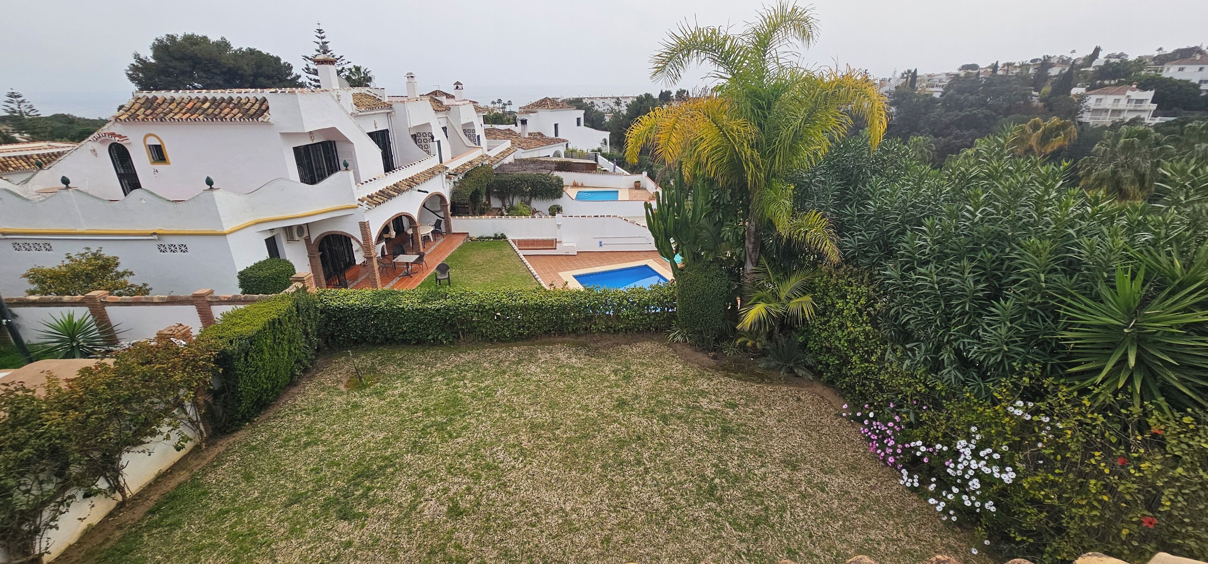 A residential property with a swimming pool, lush greenery, and a white building with a tiled roof.