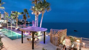 Zwembad van een resort met een rieten gazebo, palmbomen en uitzicht op de oceaan.