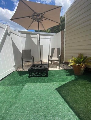 Un patio con césped artificial, una mesa y sillas, una sombrilla grande y una planta en maceta.