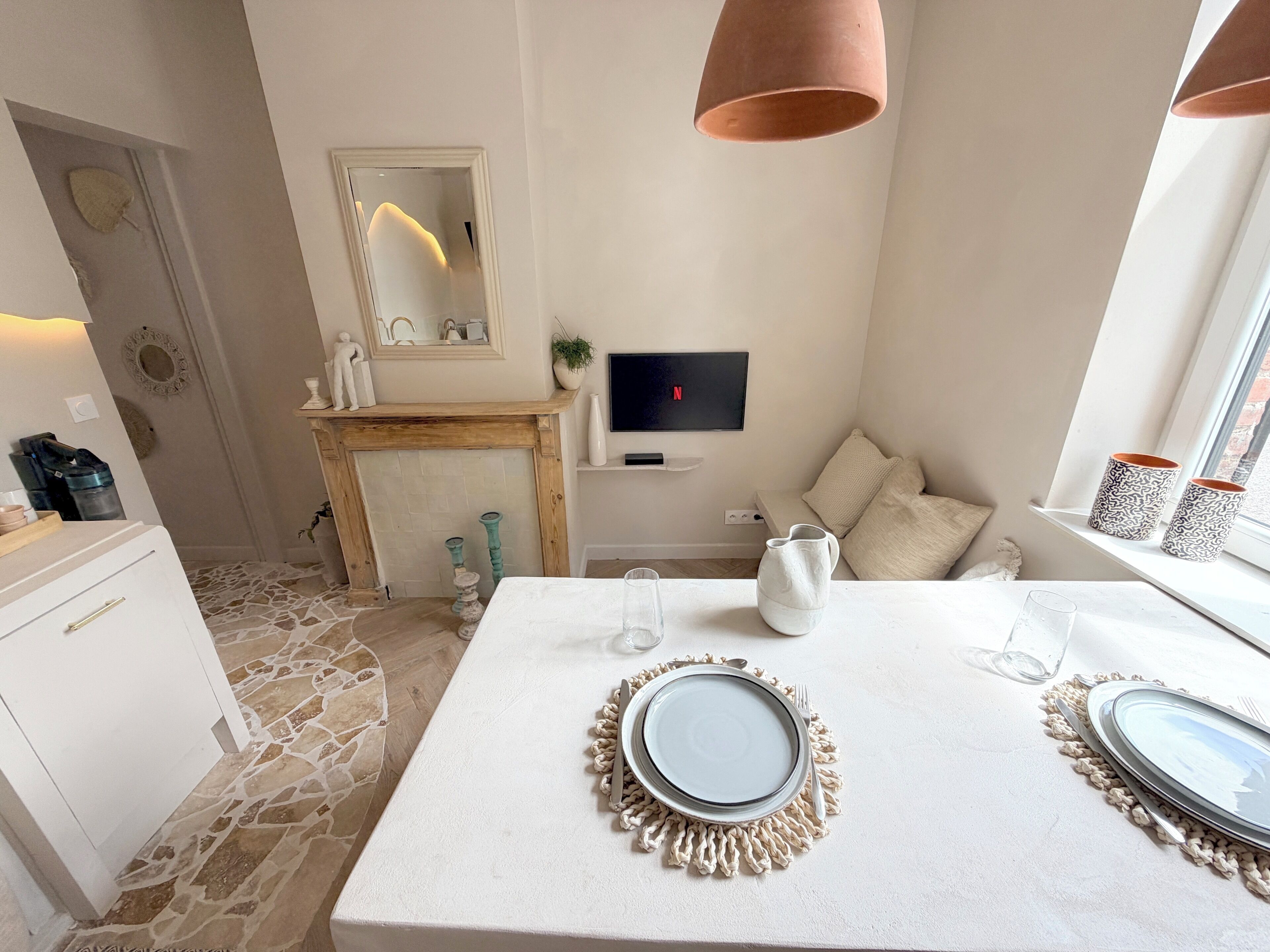 Un comedor moderno con suelo de piedra, una mesa blanca con platos, un televisor montado en la pared y una ventana con cortinas estampadas.