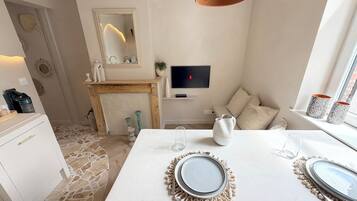 Un comedor moderno con suelo de piedra, una mesa blanca con platos, un televisor montado en la pared y una ventana con cortinas estampadas.