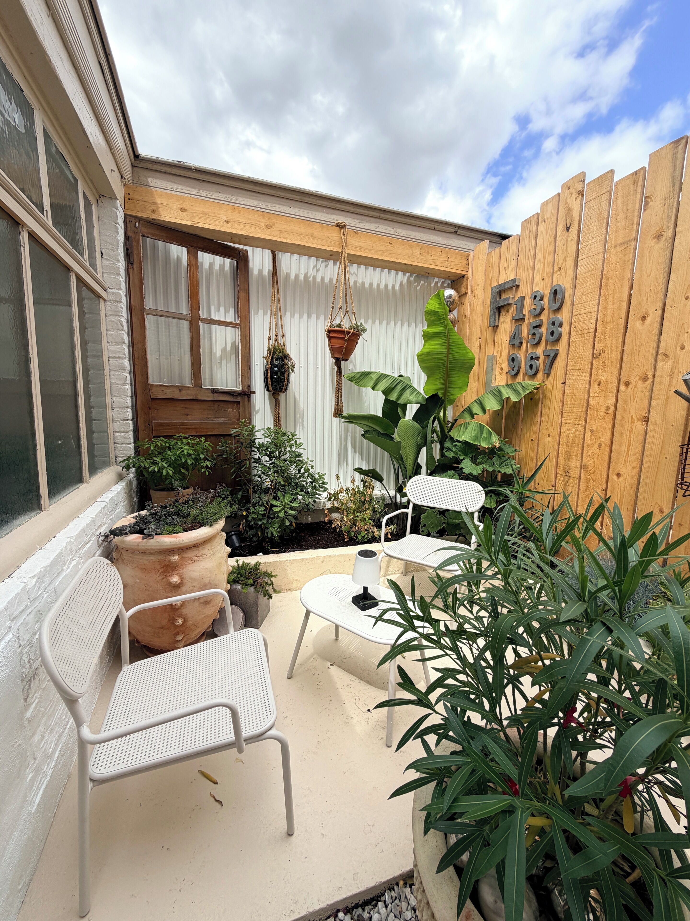 Un patio con sillas de metal blanco y una mesa, rodeado de plantas en macetas y una valla de madera.