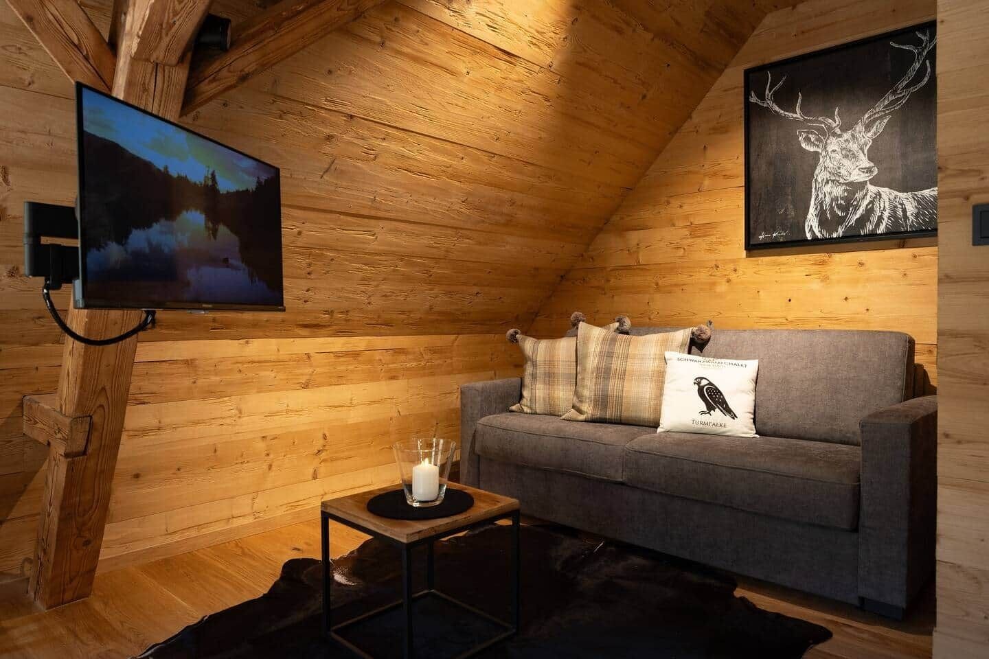 L’intérieur chaleureux d’un chalet avec un plafond en bois, un canapé gris agrémenté de coussins décoratifs, un téléviseur fixé au mur et un tableau représentant des animaux sauvages, encadré.