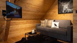 L’intérieur chaleureux d’un chalet avec un plafond en bois, un canapé gris agrémenté de coussins décoratifs, un téléviseur fixé au mur et un tableau représentant des animaux sauvages, encadré.