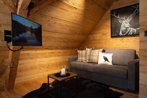 A cozy cabin interior with a wooden ceiling, a gray sofa with decorative pillows, a mounted television, and a framed wildlife painting. - Schwarzwald Chalet -Wilde Rench- “Turmfalke“ (Bad Peterstal-Griesbach)
