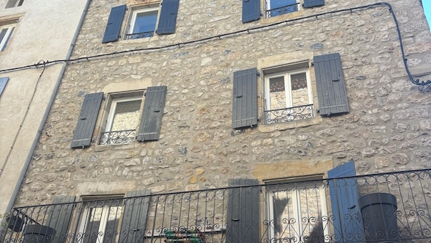 Un immeuble à plusieurs étages, doté d’une façade en pierre, de nombreuses fenêtres avec volets et d’un balcon orné d  ’une rambarde en métal.