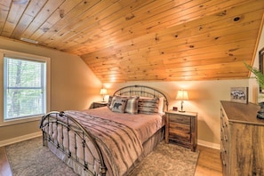 A bedroom with a sloped ceiling, a bed with a headboard, bedside tables, a dresser, and a window with blinds. - Blue Ridge Pkwy | Hot Tub + Fire Pit | EV Charger (Watauga County)