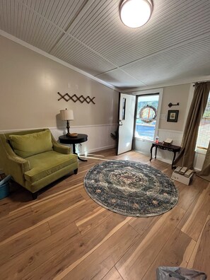Een woonkamer met een groene bank, een rond tapijt, houten vloeren, een lamp en een deur naar buiten.