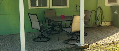 Ein überdachter Patio-Bereich mit Tisch, Stühlen, einem Mülleimer und einem Gartenschlauch.