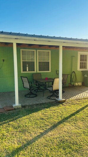 Ein überdachter Patio-Bereich mit Tisch, Stühlen, einem Mülleimer und einem Gartenschlauch.
