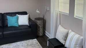 Ein Wohnzimmer mit einem schwarzen Sofa, einem kleinen schwarzen Hocker, einem weißen und türkisnen Kissen, einem weißen und türkisnen gestreiften Kissen, einer weißen und türkisnen Decke, einem weißen und türkisnen Teppich, einer weißen Wand mit einem Gemälde, einer weißen Stehlampe und einem Fenster mit Jalousien.