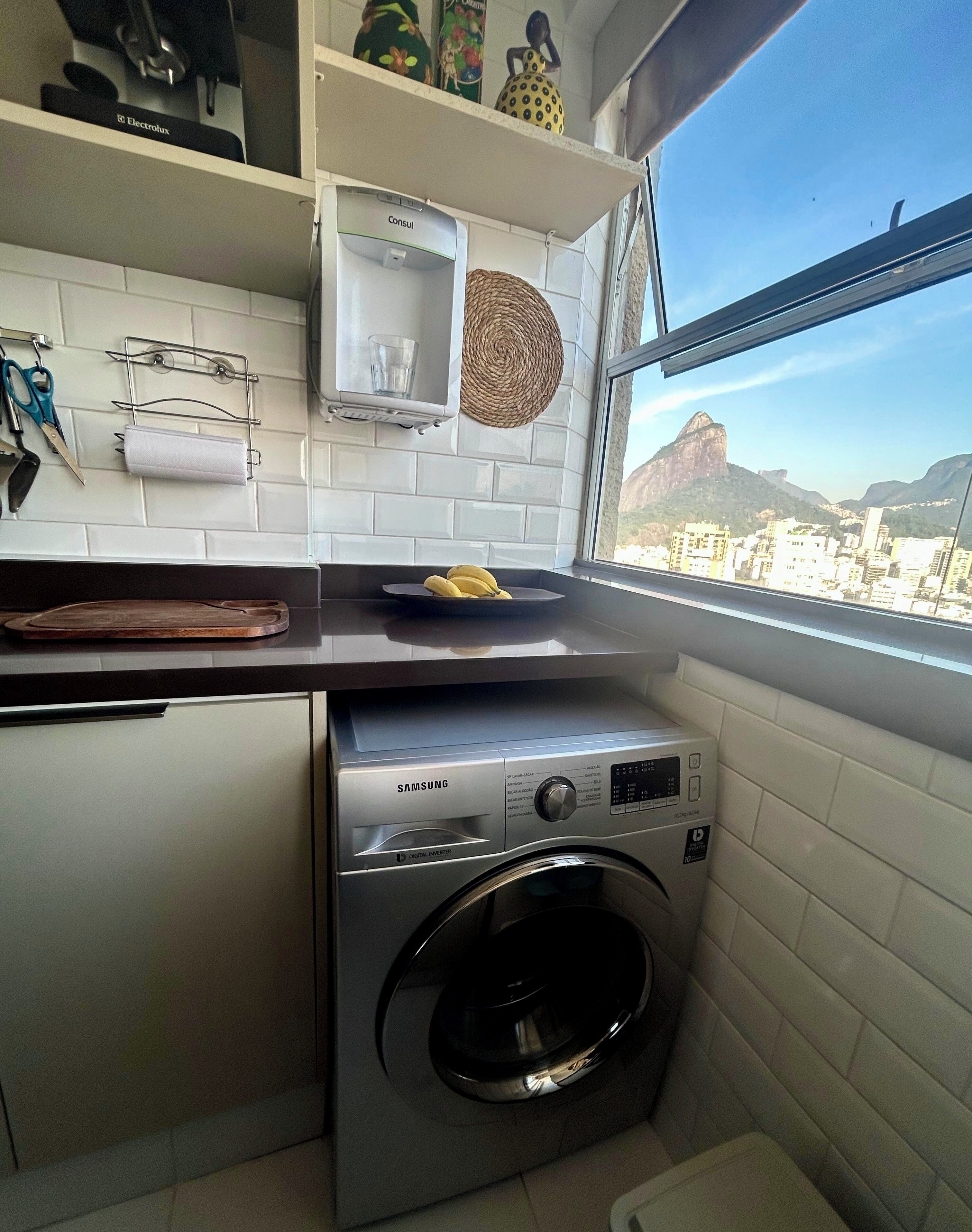 Uma cozinha compacta com máquina de lavar, janela e vista para a cidade, e prateleira com utensílios de cozinha.