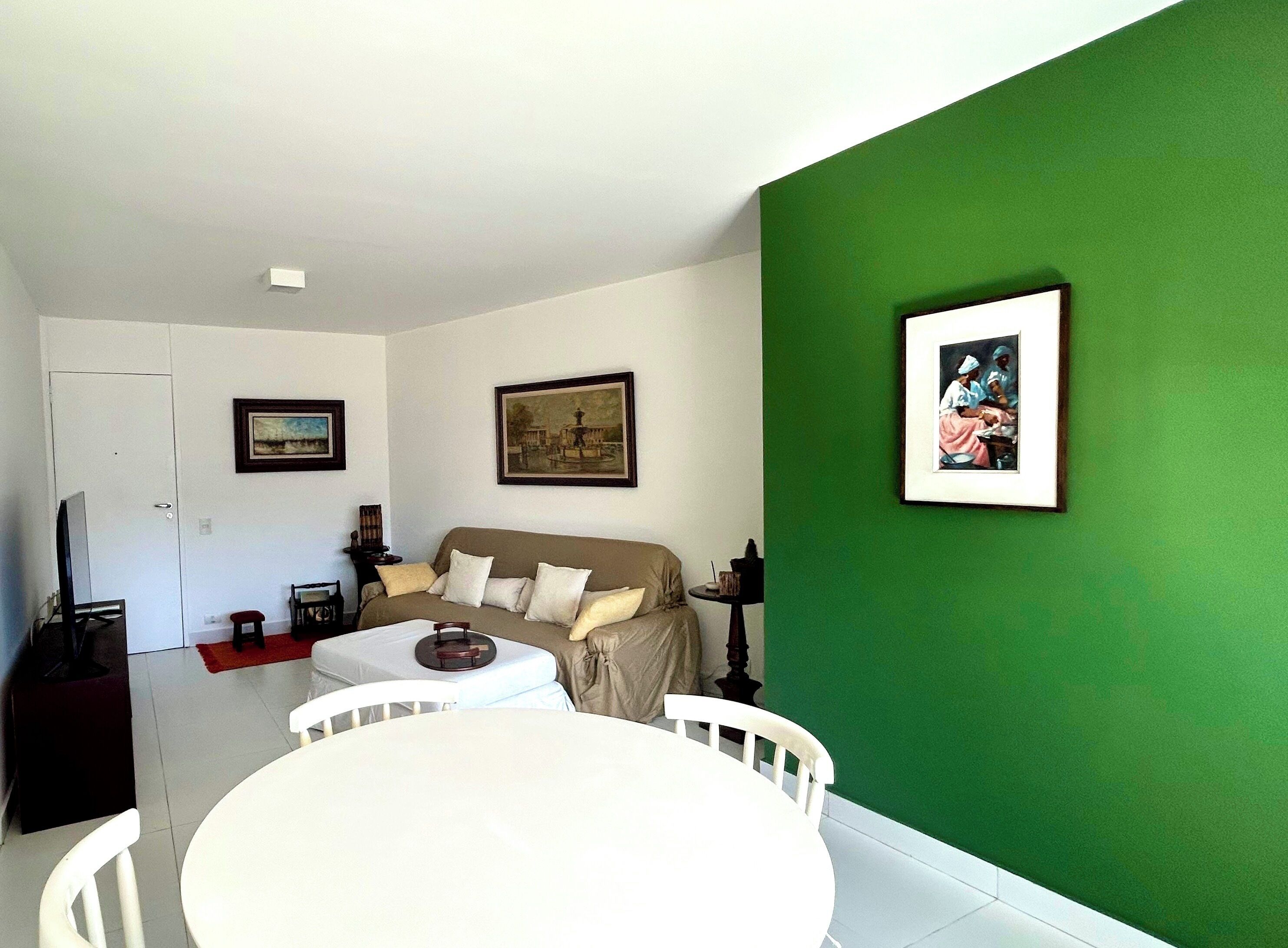 Uma sala de estar com uma parede verde de destaque, um sofá branco, uma mesa de jantar com cadeiras e obras de arte emolduradas nas paredes.