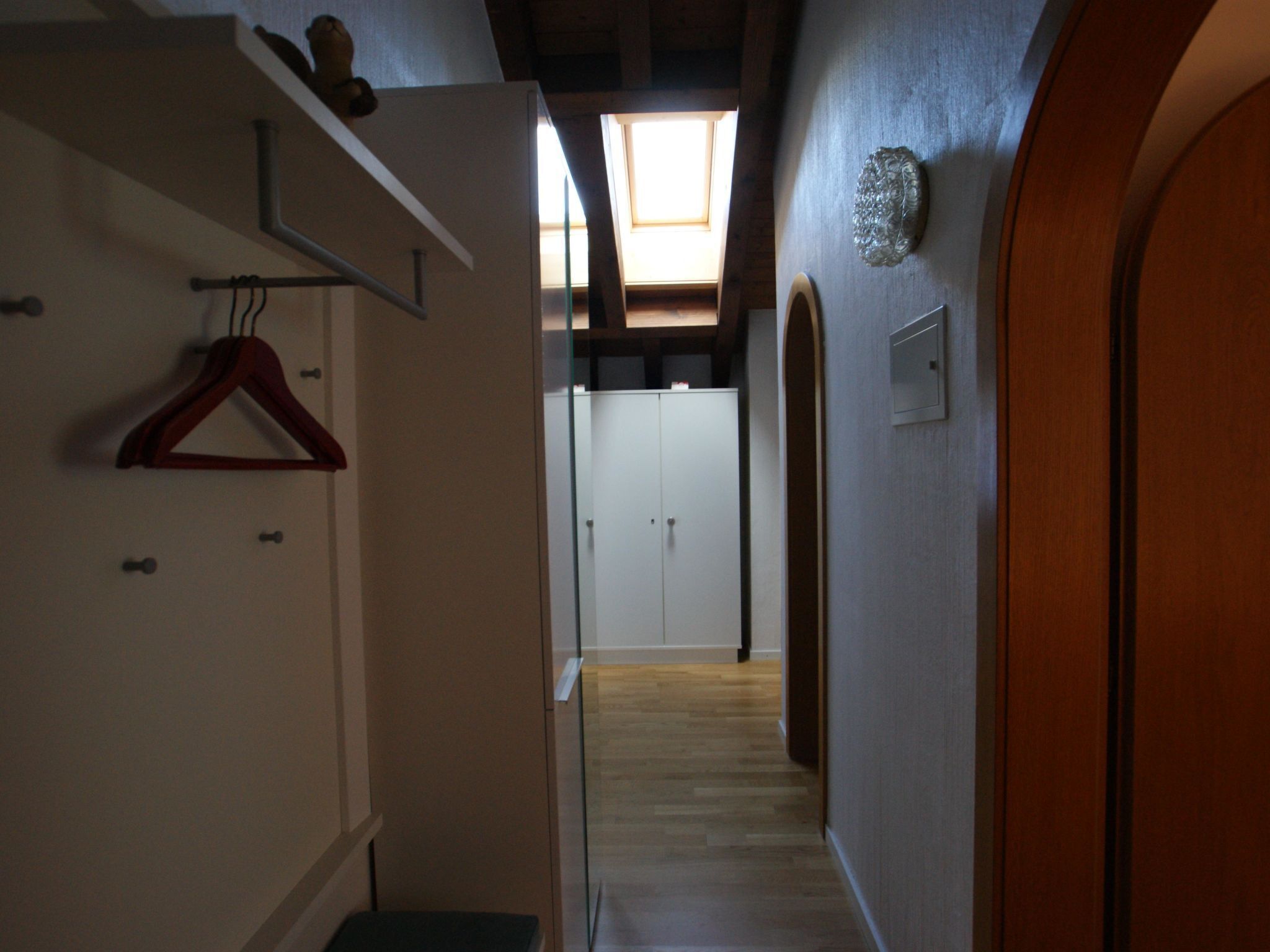 A hallway with a wooden floor, a coat rack, a door, and a wall-mounted light switch.