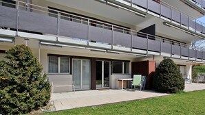 En moderne boligbygning i to etager med balkon, lille udendørs siddeområde og velplejet græsplæne.