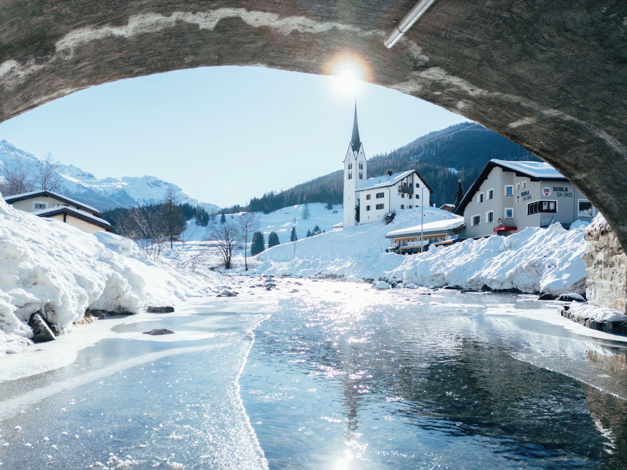 A winter scene with a frozen river under a bridge, a church, and buildings with snow-covered roofs.