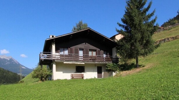 Un chalet à deux étages doté d’un balcon, situé sur une colline verdoyante, avec une montagne en arrière-plan.