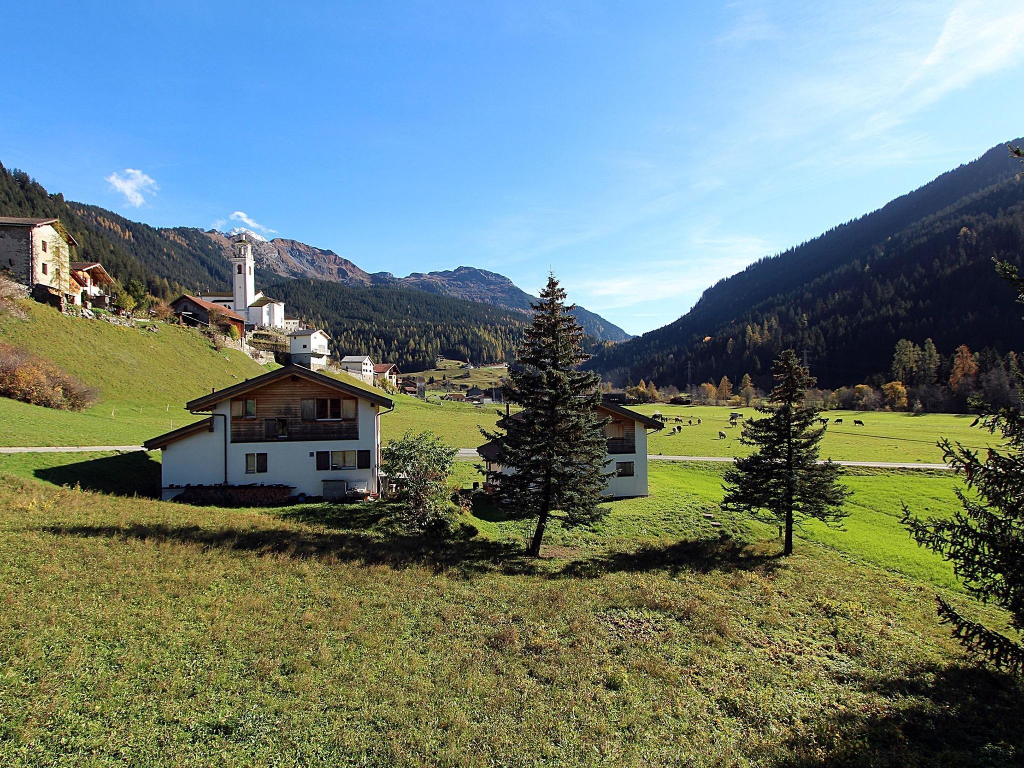 A rural landscape with a white building, a church, and mountains in the background.