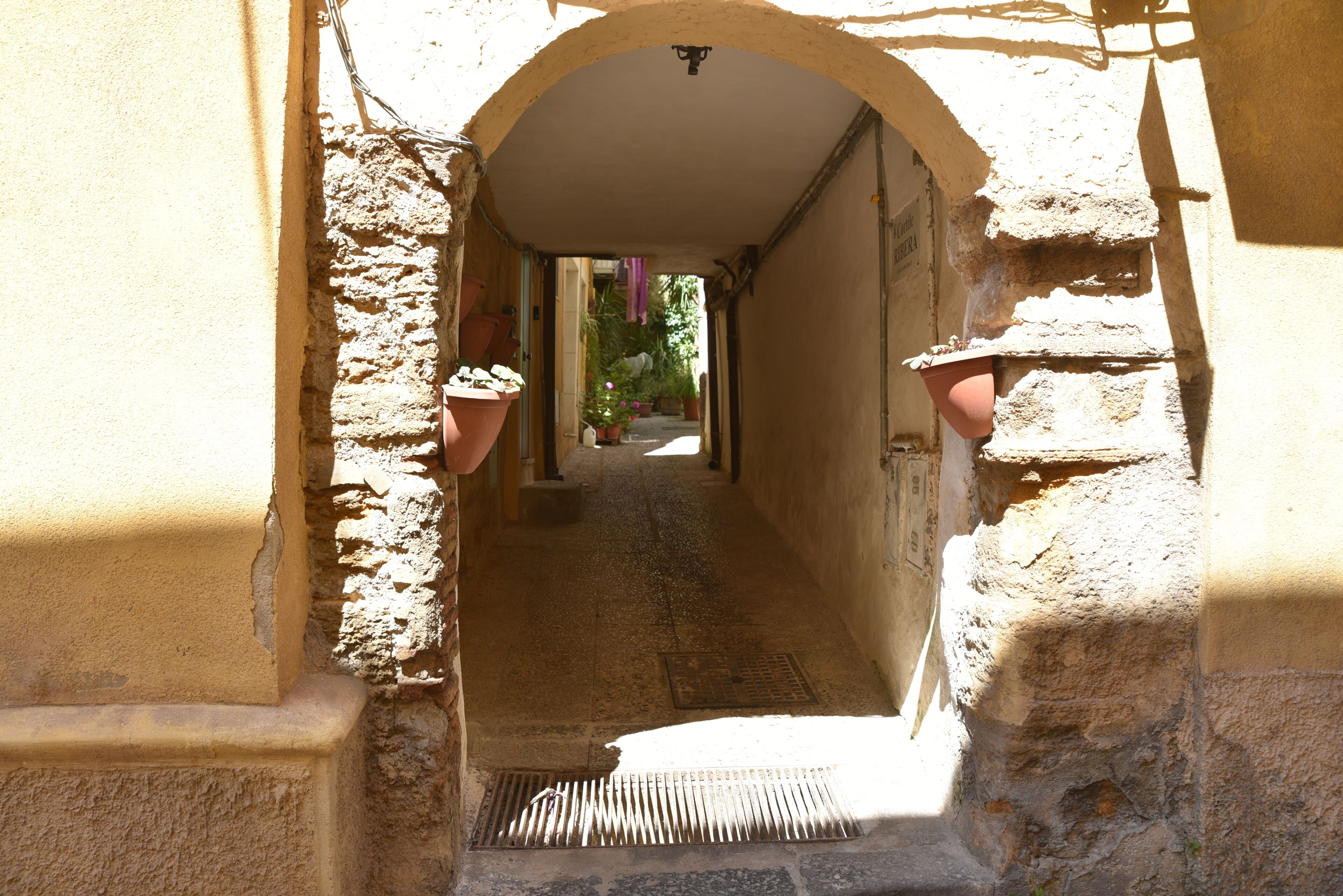 A narrow stone corridor with a metal grate at the bottom, flanked by walls with terracotta pots.