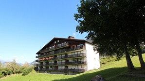 Un immeuble à plusieurs étages, doté de balcons, situé sur une pente herbeuse avec une montagne en arrière-plan.